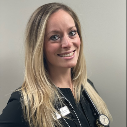 Smiling blonde female nurse wearing scrubs and stethoscope, standing against plain background.