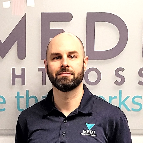 Medi Healthcare employee smiling in front of company logo, bald with beard, navy blue polo shirt